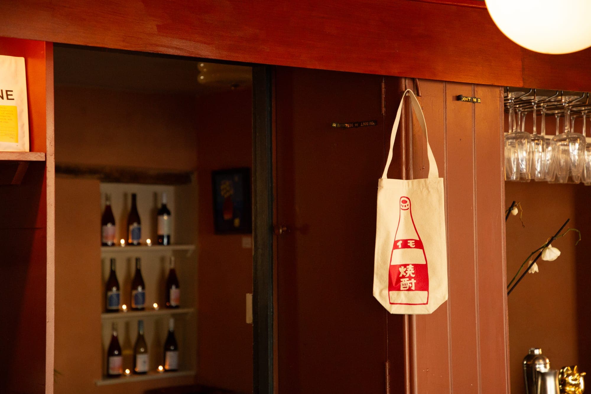 The bar: a shochu tote hanging on a door, bottles in a wall niche lit by tealights beyond, wine glasses racked on the right