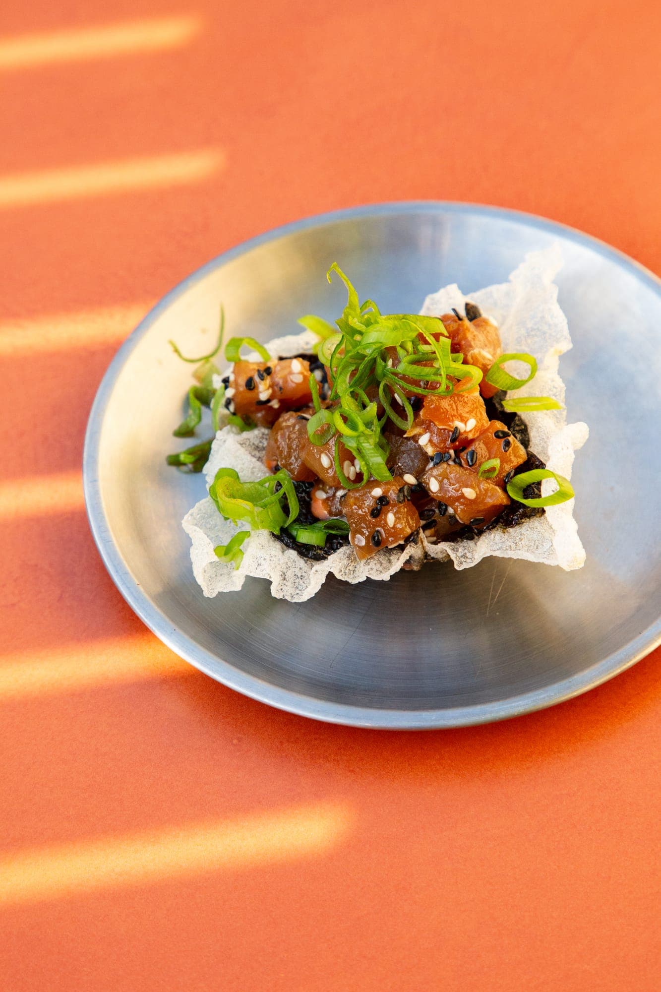 Tuna crudo with spring onion and sesame on a rice cracker, on a steel plate, window light striping the orange table