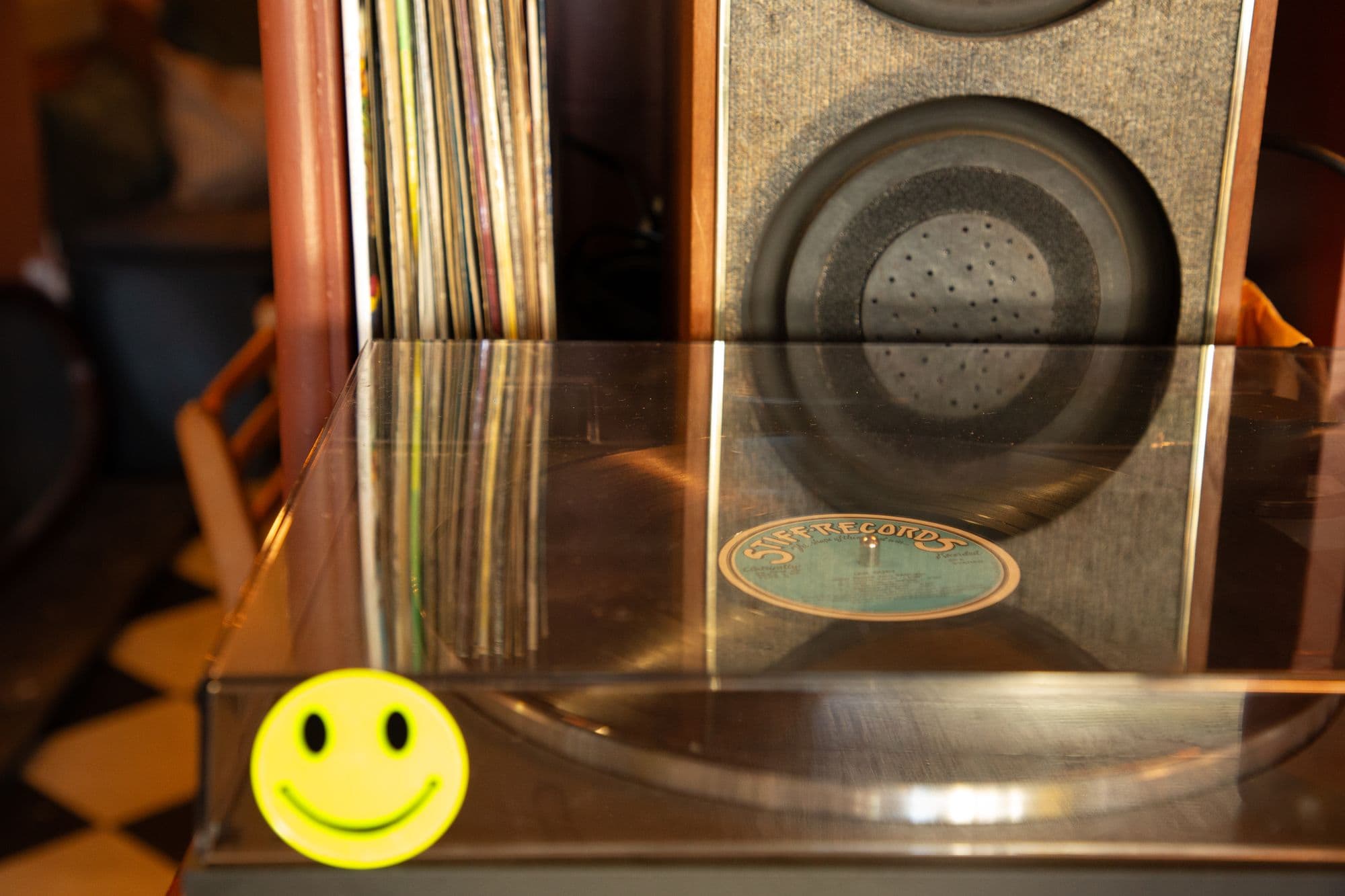 A turntable with a smiley sticker on its lid, record sleeves and a speaker behind it