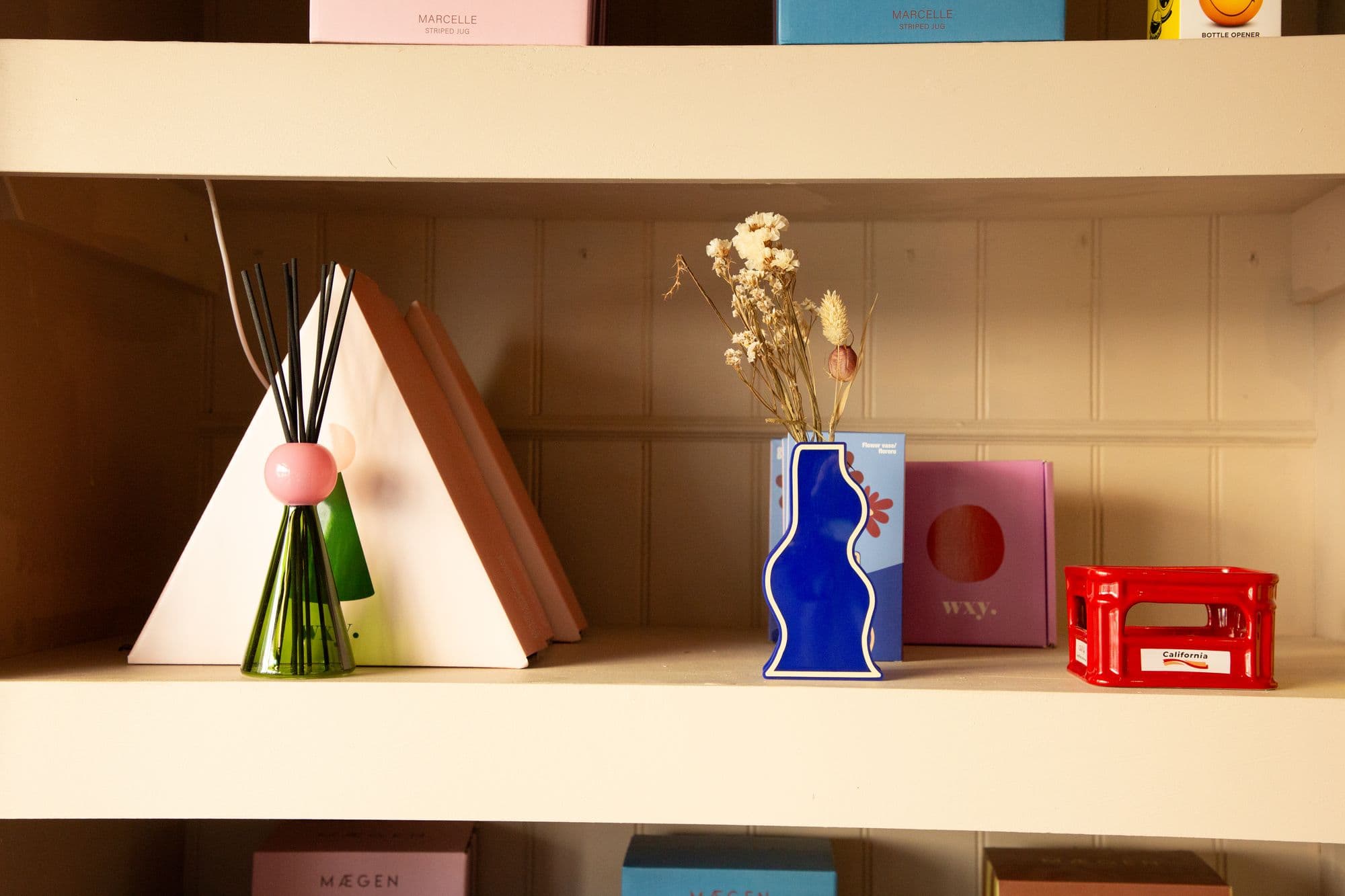 A shop shelf holding a reed diffuser, a wavy blue vase of dried flowers and a red crate