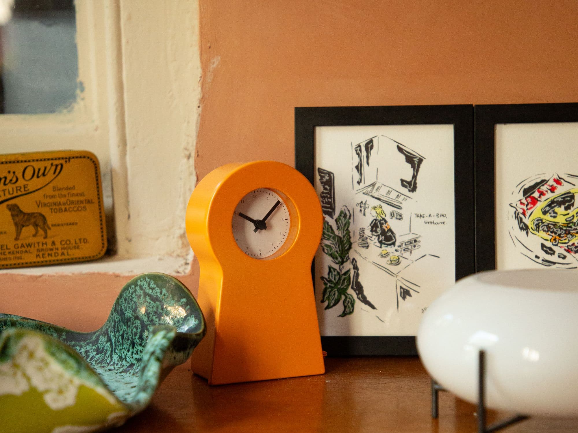 An orange clock, a green ceramic bird and framed drawings on a wooden sill against a pink wall