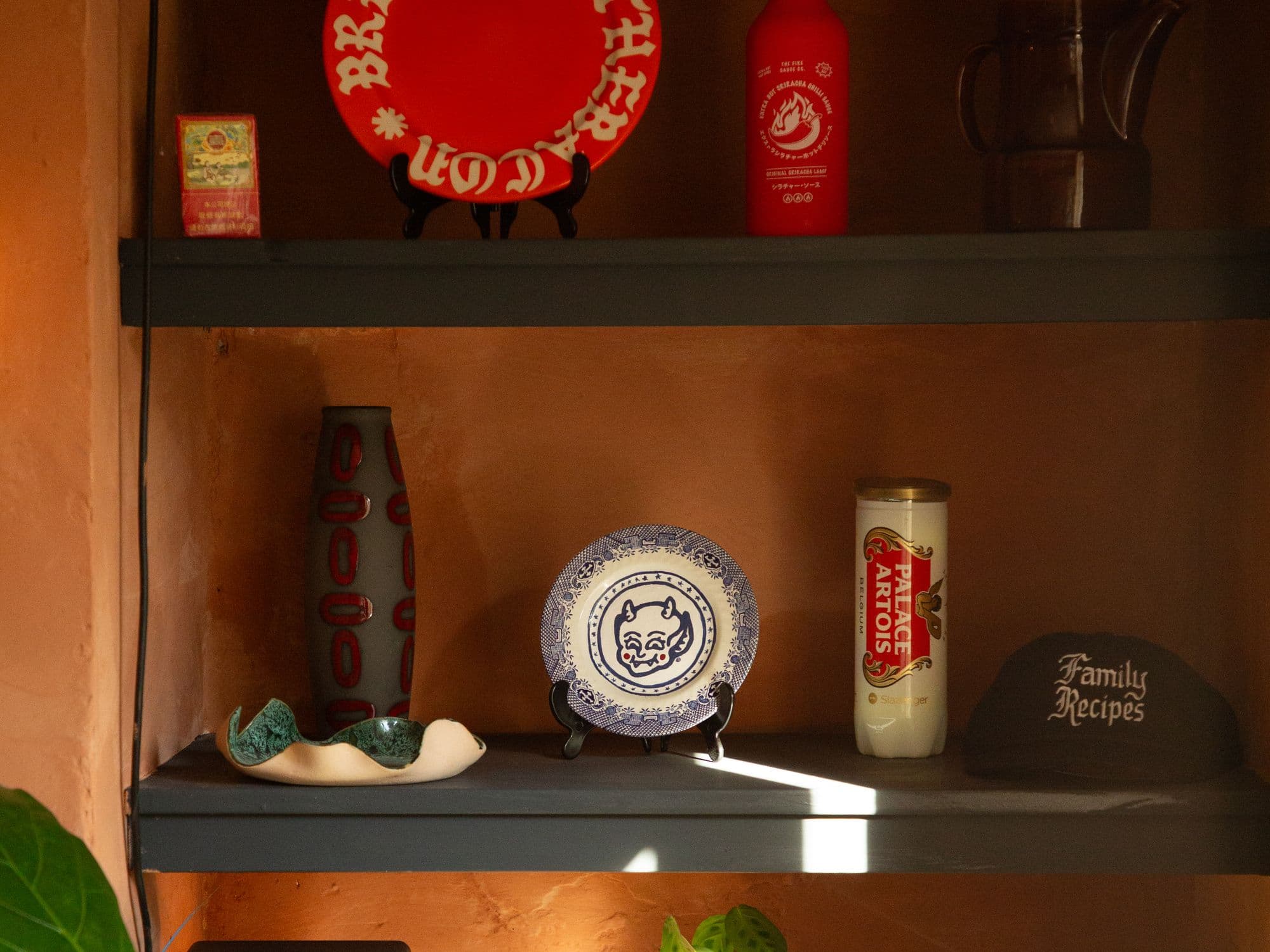 Painted shelves on a terracotta wall holding a red plate lettered in gothic script, a bottle of hot sauce and a patterned plate