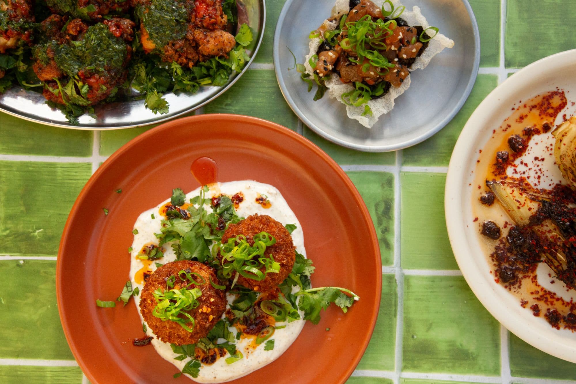 Four dishes from above on green tiles: croquettes on a terracotta plate, tuna crudo on a rice cracker, fried chicken with green sauce and charred cabbage